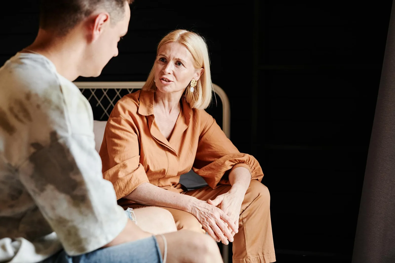 A mother and her adult son sitting together in warm indoor light, having a quiet, focused conversation.
