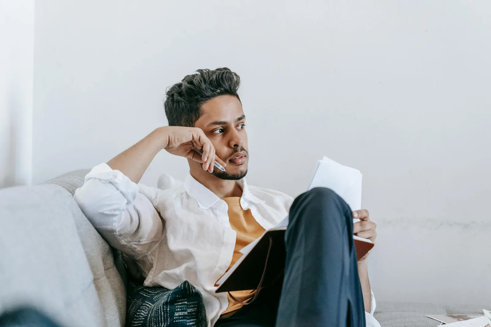 A young adult sitting on a couch with a planner open, looking away in thought in soft natural light.
